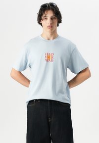 Lichtblauwe katoenen t-shirt met een rood-oranje tekstgrafiek "FEED YOUR MIND" op de borst, gecombineerd met een donkere spijkerbroek.