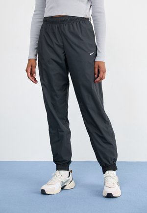 Persoon die zwarte Nike joggingbroek en witte Nike sneakers draagt en op een blauwe vloer staat tegen een effen witte achtergrond.
