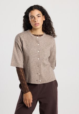 Jonge vrouw met krullend haar, gekleed in een beige gebuttonde cardigan over een bruin kanten longsleeve shirt en donkere broek tegen een effen achtergrond.