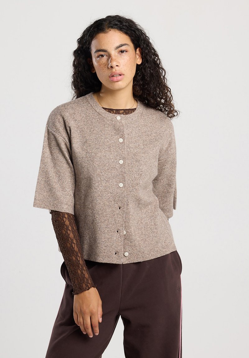 Nuori nainen, jolla on kihara tukka, pukeutuneena beigeen napitettuun neuleeseen, ruskean pitsisen pitkähihaisen paidan ja tummien housujen kanssa yksinkertaista taustaa vasten.