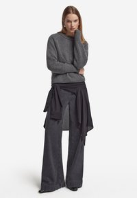 Maglione grigio lavorato a maglia con polsini a coste, abbinato a pantaloni neri di denim a gamba larga e a vita alta. Camicia nera a strati annodata in vita.