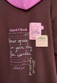 Hnědá mikina s růžovou kapucí, která představuje potištěné grafiky objednávek pro hosty, zprávy a ručně psaný text v různých barevných inkotech.