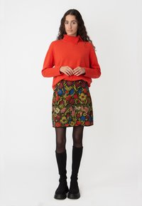 Oranje trui met hoge kraag, gecombineerd met een minirok met een bloemenpatroon met meerkleurige bloemen tegen een donkere achtergrond.