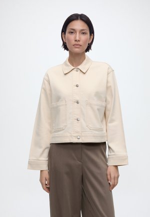 Frau mit glatten dunklen Haaren, die eine kremfarbene Jacke mit Knöpfen und lockere braune Hosen trägt, vor einem einfarbigen hellen Hintergrund.