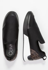 Czarne buty wsuwane z gładkim wykończeniem, zdobione teksturowanym brązowym wzorem i białą podeszwą. Materiał wydaje się elastyczny i lekki.