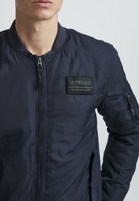 Chaqueta bomber azul marino con cierre delantero, cuello de canalé y bolsillo en la manga izquierda. Presenta un parche de tela negra con "11 PROJECT" y el año de establecimiento.