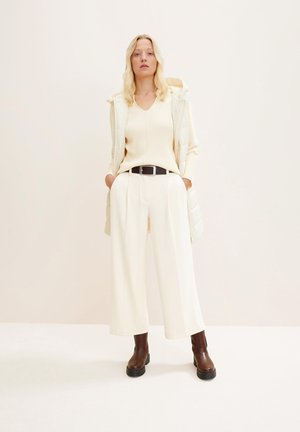 Maglione a coste con scollo a V di colore crema, gilet imbottito beige chiaro e pantaloni larghi di colore crema. Stivali alla caviglia marroni con suola spessa. Cintura nera in evidenza.