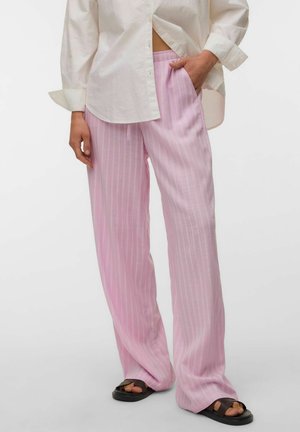Personne portant un pantalon ample rayé rose clair avec des poches, une chemise blanche à manches longues avec poignets, et des sandales noires à enfiler, debout sur fond blanc.