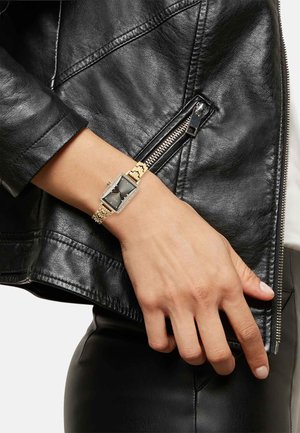 Guldur med rektangulært ansigt, sort urskive og krystalaccenter på en kædearmbånd, der bæres på en hånd mod en sort læderjakke.