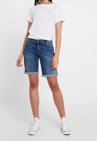 Nainen, jolla on ylös käärityt siniset farkkushortsit, yksinkertainen valkoinen t-paita ja valkoiset korkeavartiset lenkkarit, seisoo valkoista taustaa vasten.