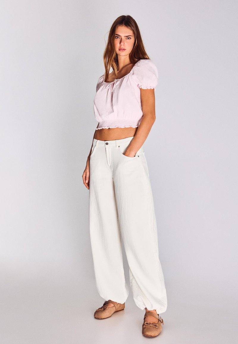 Jeune femme debout, une main dans la poche, portant un crop top à manches bouffantes rose clair, un pantalon blanc à jambes larges et des ballerines marron.