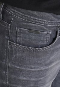 Jeans de mezclilla gris con un acabado ligeramente desgastado, que presentan un bolsillo trasero y una pequeña etiqueta negra con el nombre de la marca.