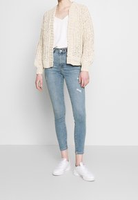 Cárdigan de punto grueso en color crema, con bordes acanalados, superpuesto sobre una blusa ligera y jeans ajustados de color azul claro con desgastes, combinados con zapatillas blancas.
