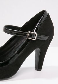 Černé semišové lodičky Mary Jane na vysokém podpatku s lakovaným koženým páskem a stříbrnou sponou, umístěné na plain světlem pozadí.