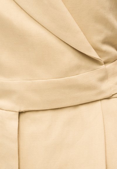 Κοντινή λήψη μπεζ υφάσματος με διπλωμένες και ραμμένες λεπτομέρειες, που δείχνει προσαρμοσμένα ράμματα και υφή.
