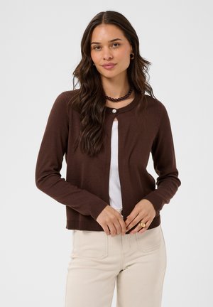 Femme aux longs cheveux foncés portant un cardigan marron par-dessus un haut blanc, un pantalon beige, un collier de perles et des boucles d'oreilles dorées en forme d'anneau.