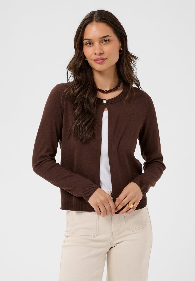 Femme aux longs cheveux foncés portant un cardigan marron par-dessus un haut blanc, un pantalon beige, un collier de perles et des boucles d'oreilles dorées en forme d'anneau.