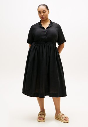 Γυναίκα που στέκεται φορώντας μαύρο φόρεμα μέσης μήκους με κουμπιά, κοντά μανίκια και μπεζ σανδάλια ανοιχτών δαχτύλων σε λευκό φόντο.