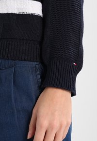 Pull en maille marine avec détail de rayure blanche ; design texturé et poignets côtelés. Petit drapeau en accent sur la manche. Porté avec un denim foncé.
