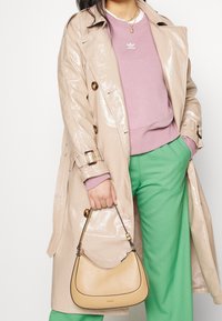 Beige lacktrenchcoat med stora knappar, kombinerad med en rosa sweatshirt och gröna byxor. Håller en beige läderaxelväska med svarta accenter.