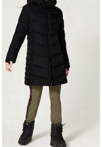 Abrigo acolchado negro con diseño de acolchado horizontal, cremallera frontal y capucha. Combinado con pantalones verde oliva con una franja lateral y botas negras.