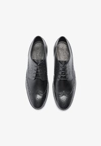 Chaussures de ville en cuir noir avec détails en brogue, bout rond, lacées et texture lisse. Logo interne visible sur les semelles intérieures.