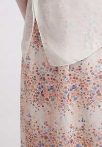 Kukkakuvioinen läpikuultava pusero kermanvärisenä, jossa oransseja, sinisiä ja vaaleanpunaisia pieniä kukkia. Kerroksellinen malli, sileä pinta ja kaareva helma.