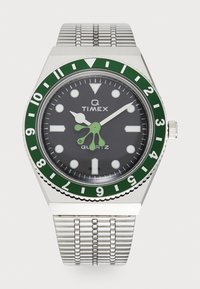 Montre-bracelet Timex avec cadran noir, lunette verte et blanche, bracelet en métal argenté et aiguille des heures unique de couleur verte avec trois extrémités arrondies.