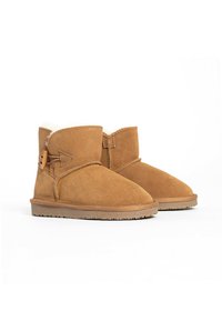Botas de tobillo de ante marrón con un diseño de solapa, forradas con suave piel de pelo, y un detalle decorativo de botón en el costado. Suela de goma plana.
