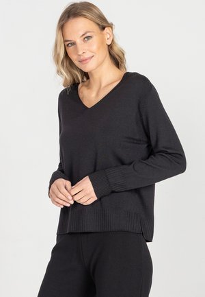 Blondynka o jasnych oczach, ubrana w czarny sweter z dekoltem w serek i czarne spodnie, stoi na tle jednolitego jasnego tła.