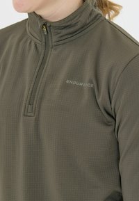 Olivgrön pullover med hög krage, zip-stängning och texturerad yta. Har "ENDURAICE"-loggan i ljusgrått på framsidan.
