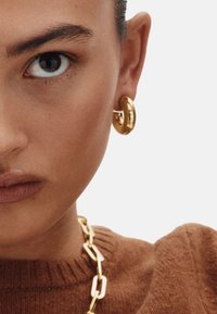 Pendientes de aro dorados y un collar de cadena gruesa dorada complementan un suéter marrón de punto con detalles acanalados. La piel tiene un acabado natural.