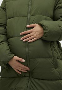 Zelena puhovka s quiltasto teksturo in zapiranjem na zadrgo. Roka modela počiva na zaobljeni sredini telesa, kar poudarja prileganje jakne.
