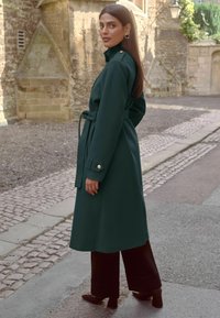 Tamnozeleni plašt s remenom, ovratnikom, bočnim džepovima i manžetama na dugmad, uparen s širokim crnim hlačama i cipelama s petom.