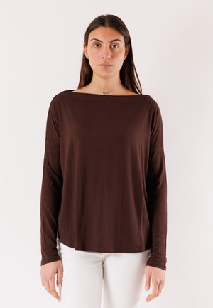 Donna che indossa una maglia marrone a maniche lunghe e ampie con scollo a barca e pantaloni bianchi, in piedi contro uno sfondo chiaro e uniforme.