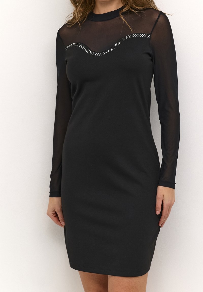 Femme portant une robe noire près du corps avec des manches longues transparentes et un col à festons à pois.