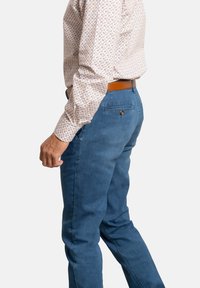 Camisa beige con un patrón ligero y de mangas largas, combinada con pantalones de mezclilla azules y un cinturón de cuero marrón. Vista lateral que muestra el ajuste y la textura.