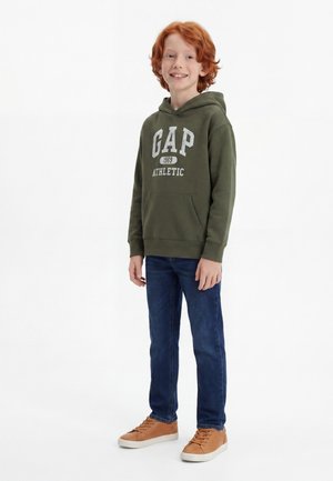 Κοκκινομάλλο αγόρι που χαμογελά, φοράει μια χακί φούτερ GAP Athletic, μπλε τζιν και μπεζ αθλητικά παπούτσια, στέκεται μπροστά σε λευκό φόντο.