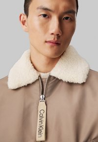 Cazadora bomber marrón con cuello de borreguito crema, cremallera plateada y una etiqueta que dice "Calvin Klein". Textura exterior suave.