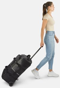 Schwarze Rollreisetasche mit einem stabilen Griff, mehreren Reißverschlusstaschen und seitlichen Riemen, die von einer Person in hellen Jeans gezogen wird.