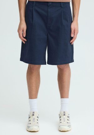 Mężczyzna ubrany w granatowe spodenki do kolan, białe skarpety za kostkę i białe buty sportowe, stojący na jednolitym tle.