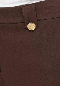 Gros plan sur un pantalon en tissu marron avec un bouton rond doré texturé sécurisant une boucle de ceinture à la taille.
