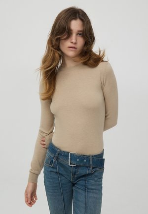 Maglietta beige a maniche lunghe con scollatura rotonda, design aderente e polsini a coste, abbinata a jeans azzurri a vita alta con una cintura larga.