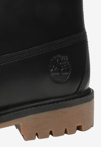 Botín de cuero negro con una superficie texturizada, que cuenta con un prominente logotipo de árbol y una suela gruesa color beige con ranuras para mayor agarre.
