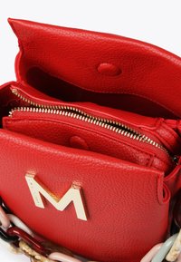 Červená kožená kabelka s zlatým logem "M", s texturovaným povrchem, kapsou na zip a dekorativním řetízkovým popruhem v různých barvách.