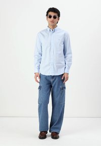 Camisa de botones azul claro, pantalones cargo de mezclilla con bolsillos y zapatos marrones. Los accesorios incluyen gafas de sol redondas. De pie sobre un fondo neutro.