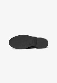 Schwarze Schuhsohle mit einer strukturierten GummOberfläche, die eine abgerundete Form und einen flachen Absatz aufweist. Beinhaltet ein kleines rechteckiges Element in der Nähe des Randes.