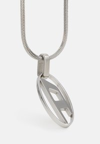 Silberne Metallkette mit einem flachen, ovalen Anhänger, der ein ausgeschnittenes geometrisches Design aufweist. Die Kette ist glatt und hat eine flexible Textur.