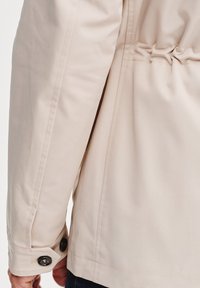 Beżowa kurtka z marszczonym tyłem, gładką fakturą i czarnymi guzikami na mankietach. Dopasowany krój z wyraźnymi szwami.