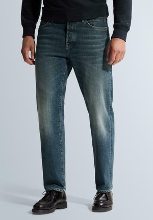 Homme portant un jean bleu délavé droit, des chaussures noires et un haut noir à manches longues, debout devant un fond uni.